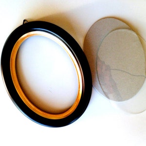 May include: A black oval picture frame with a gold inner edge. The frame is on a white surface with a clear plastic oval mat and a cardboard oval mat.