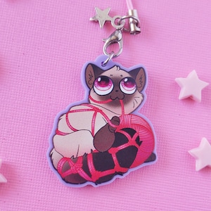 May include: A purple and white phone charm featuring a cartoon cat with pink eyes, tied up with red string and holding a ball of yarn.