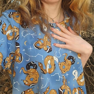 May include: A blue t-shirt with a pattern of brown snakes and constellations. The snakes are outlined in black and have white crosses on them. The constellations are made of white lines and stars.