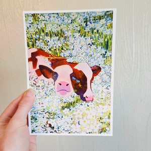 Peut inclure: Une peinture &agrave; l'aquarelle d'un veau brun et blanc couch&eacute; dans un champ de fleurs blanches. Les yeux du veau sont bleus et il regarde directement le spectateur.