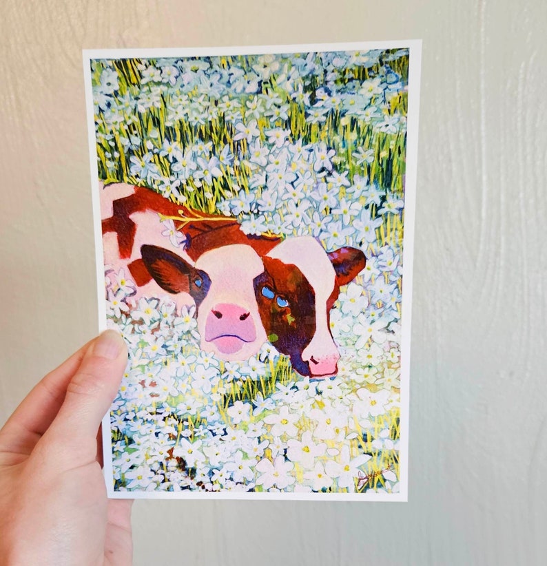 Peut inclure: Une peinture &agrave; l'aquarelle d'un veau brun et blanc couch&eacute; dans un champ de fleurs blanches. Les yeux du veau sont bleus et il regarde directement le spectateur.