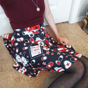 May include: A black and white skirt with a red and white print of medical symbols, hearts, stars, and a teddy bear. The skirt has a white waistband and a red and white checkered pattern on the bottom hem. The text "Alive Dead" is printed on a white rectangle on the skirt.
