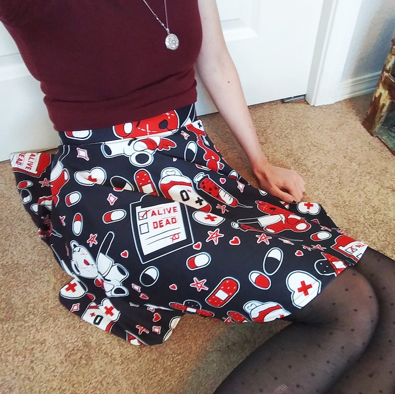 May include: A black and white skirt with a red and white print of medical symbols, hearts, stars, and a teddy bear. The skirt has a white waistband and a red and white checkered pattern on the bottom hem. The text "Alive Dead" is printed on a white rectangle on the skirt.