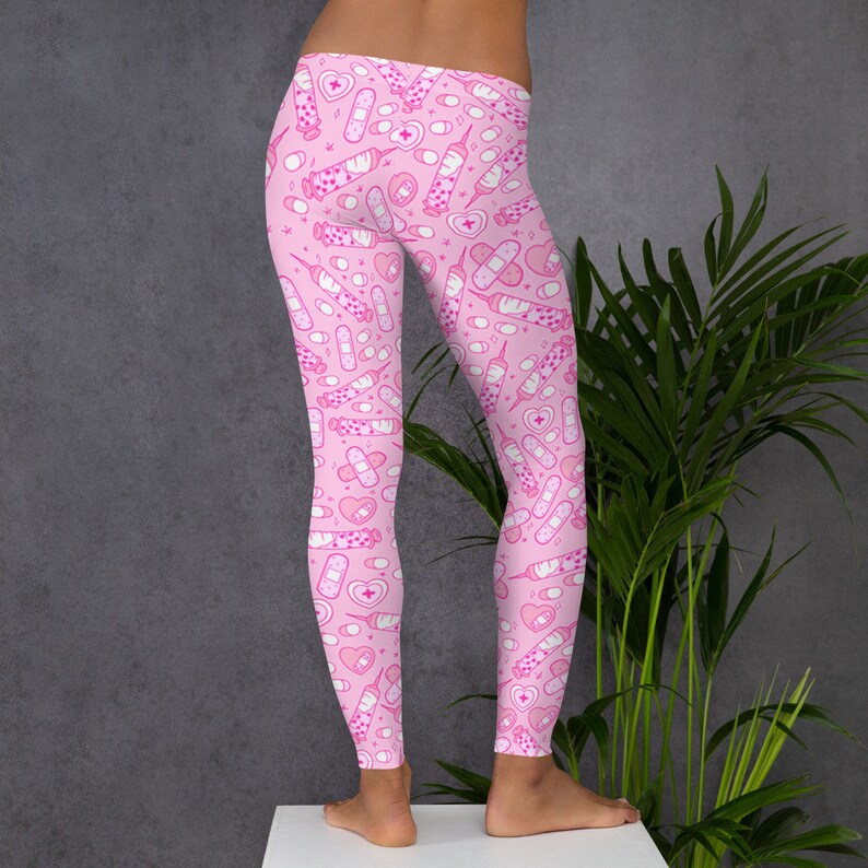 May include: Pink leggings with a repeating pattern of white medical supplies, including syringes, bandages, pills, and hearts. The leggings are made of a soft, stretchy material.