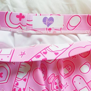 Pode incluir: Saia rosa com el&aacute;stico branco na cintura e uma etiqueta branca com o texto "fss" em um cora&ccedil;&atilde;o roxo. A saia &eacute; coberta por um padr&atilde;o de cora&ccedil;&otilde;es rosas, bandagens, p&iacute;lulas e outros s&iacute;mbolos com tema m&eacute;dico.