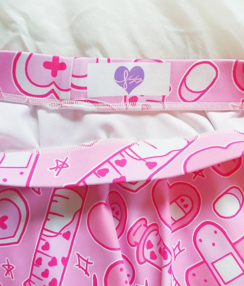 Pode incluir: Saia rosa com el&aacute;stico branco na cintura e uma etiqueta branca com o texto "fss" em um cora&ccedil;&atilde;o roxo. A saia &eacute; coberta por um padr&atilde;o de cora&ccedil;&otilde;es rosas, bandagens, p&iacute;lulas e outros s&iacute;mbolos com tema m&eacute;dico.