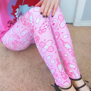 May include: Pink leggings with a repeating pattern of medical-themed illustrations, including syringes, bandages, pills, and hearts. The leggings are a light pink color with white and darker pink accents. The person is wearing black shoes.