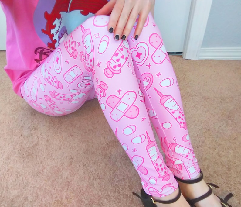 May include: Pink leggings with a repeating pattern of medical-themed illustrations, including syringes, bandages, pills, and hearts. The leggings are a light pink color with white and darker pink accents. The person is wearing black shoes.
