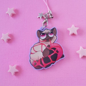 May include: A white cat with pink eyes is tied up with red string and holding a ball of red yarn. The cat is part of a charm that is attached to a silver chain with a star charm.