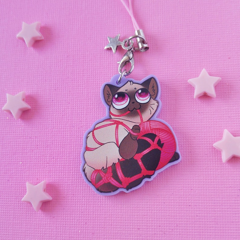 May include: A white cat with pink eyes is tied up with red string and holding a ball of red yarn. The cat is part of a charm that is attached to a silver chain with a star charm.