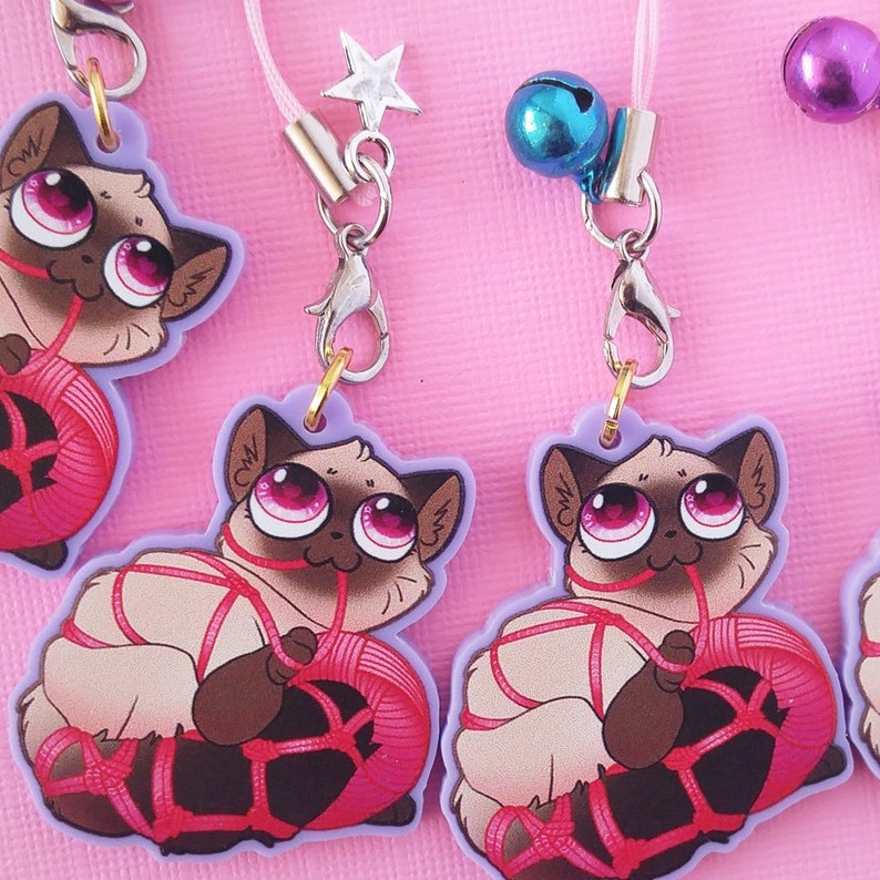 May include: Three pink and purple cat charm keychains with a pink yarn ball. Each charm features a cartoon cat with large pink eyes and a brown nose. The cats are tied up with pink string.