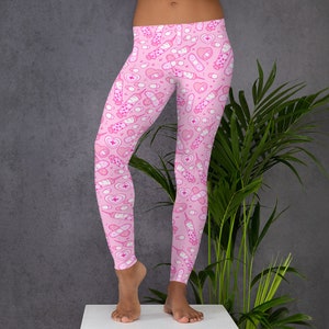 May include: Pink leggings with a repeating pattern of medical-themed illustrations. The design includes syringes, bandages, pills, and hearts. The leggings are made of a soft, stretchy material.