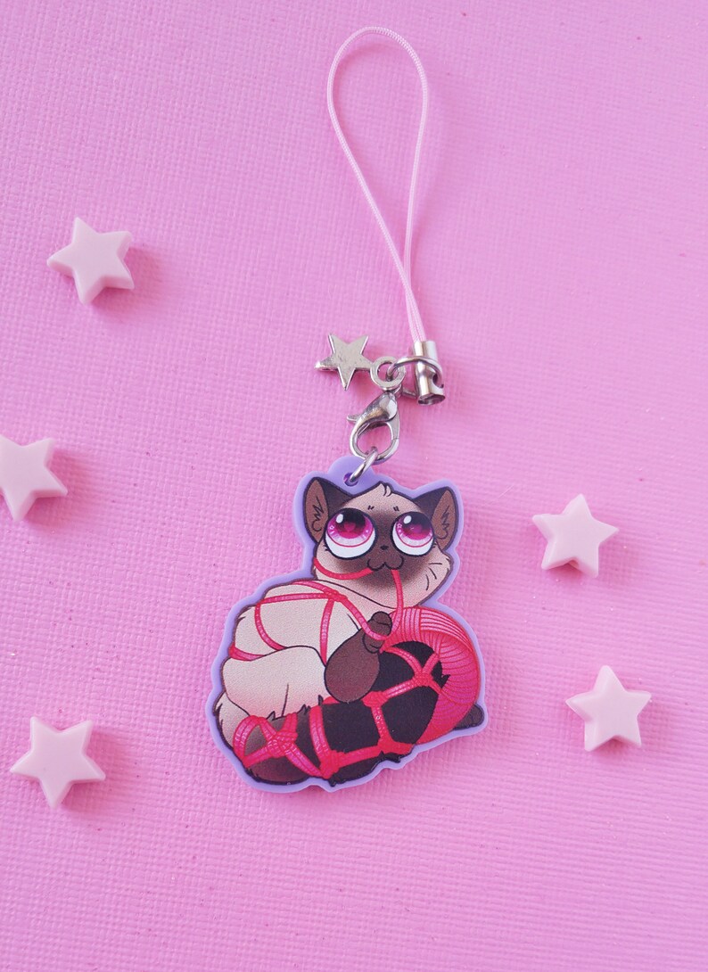 May include: A purple and white phone charm featuring a cartoon cat with pink eyes, tied up with red string and holding a ball of yarn.
