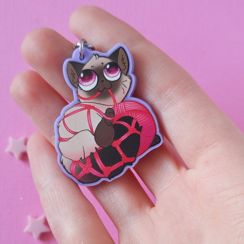 May include: A purple cat charm with pink eyes and red yarn tied around it. The cat is holding the yarn in its paws.