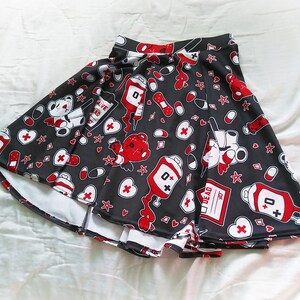 May include: A black skater skirt with a white and red print of teddy bears, hearts, bandages, pills, and blood bags. The print includes the text "Alive Dead" and "0+".