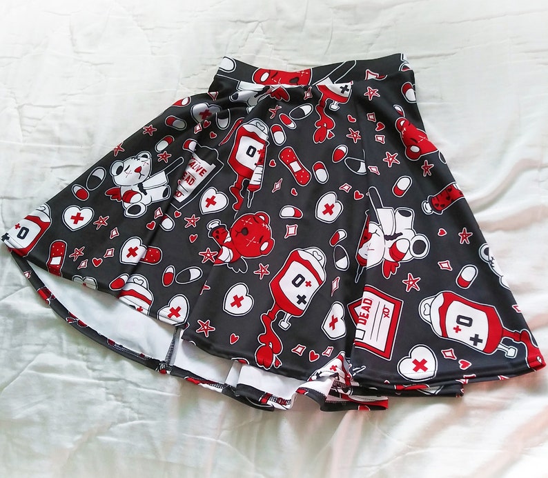 May include: A black skater skirt with a white and red print of teddy bears, hearts, bandages, pills, and blood bags. The print includes the text "Alive Dead" and "0+".