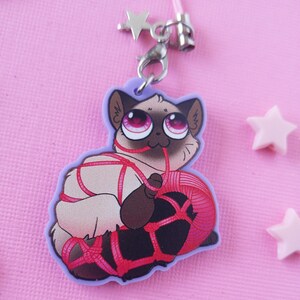 May include: A white cat with pink eyes is tied up with red string and holding a ball of red yarn. The cat is a charm on a purple background.