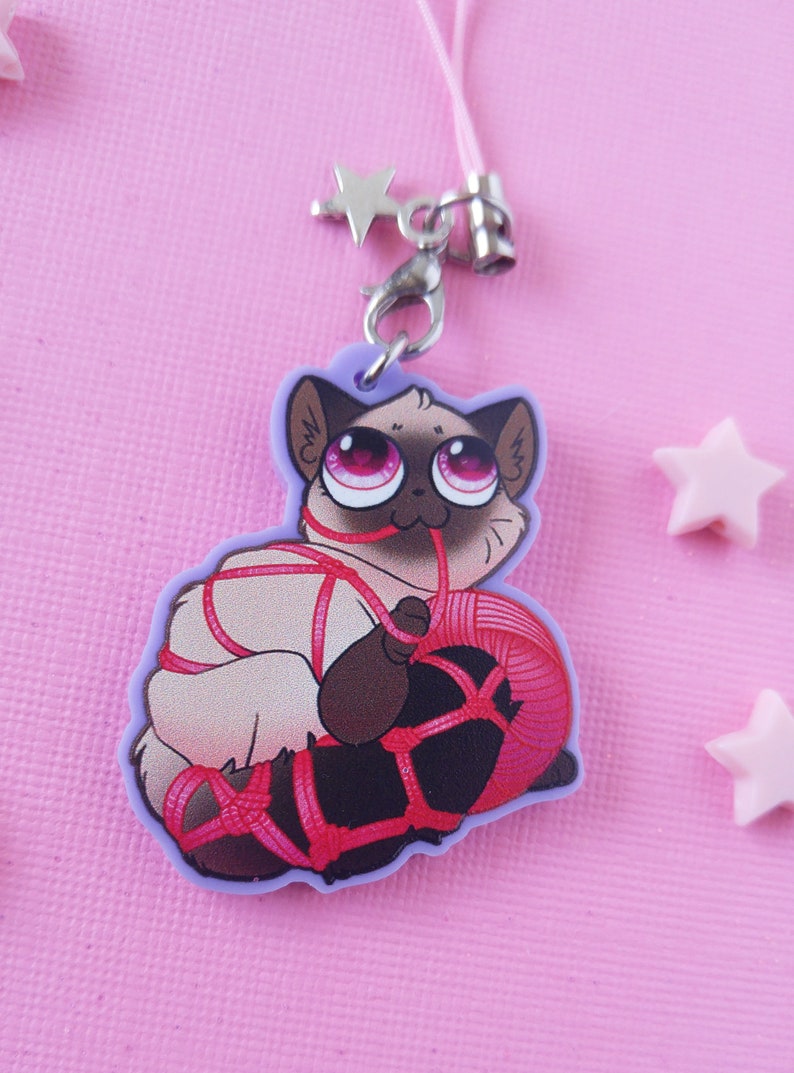 May include: A white cat with pink eyes is tied up with red string and holding a ball of red yarn. The cat is a charm on a purple background.