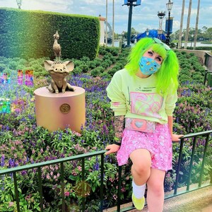 Pode incluir: Uma pessoa com cabelo verde brilhante vestindo uma camisa de l&atilde; amarela clara com um design gr&aacute;fico rosa e branco, uma saia rosa e branca e uma bolsa rosa em forma de cora&ccedil;&atilde;o. A pessoa est&aacute; em p&eacute; na frente de uma est&aacute;tua de bronze de um porco e um p&aacute;ssaro.