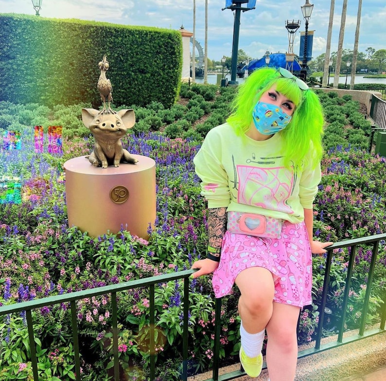 Pode incluir: Uma pessoa com cabelo verde brilhante vestindo uma camisa de l&atilde; amarela clara com um design gr&aacute;fico rosa e branco, uma saia rosa e branca e uma bolsa rosa em forma de cora&ccedil;&atilde;o. A pessoa est&aacute; em p&eacute; na frente de uma est&aacute;tua de bronze de um porco e um p&aacute;ssaro.