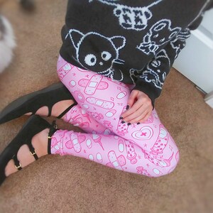 May include: A person wearing a black sweater with white cartoon graphics and pink leggings patterned with medical-themed illustrations. The person is also wearing black flats with gold buckles.