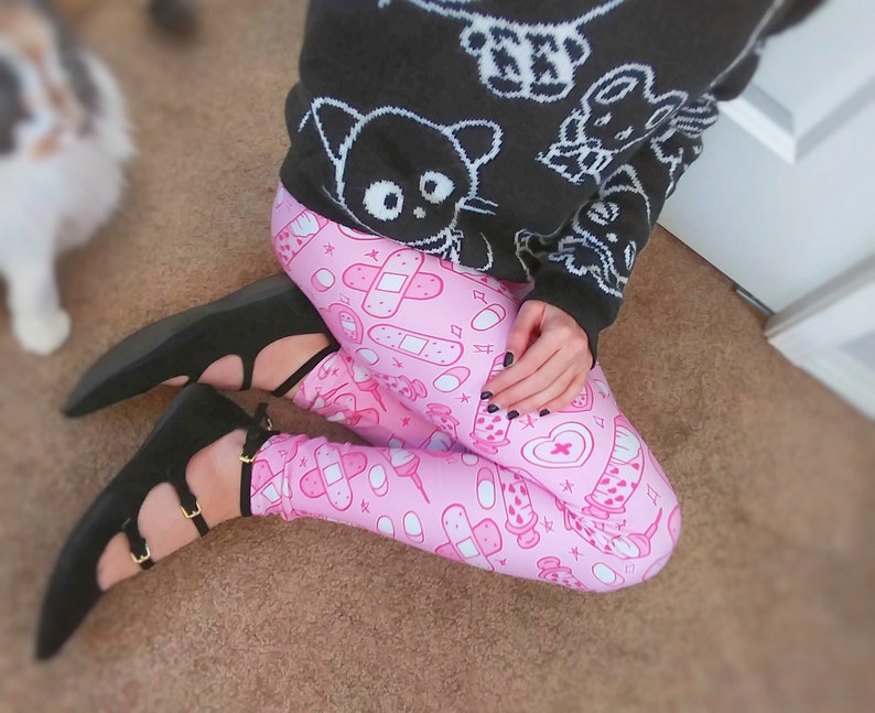 May include: A person wearing a black sweater with white cartoon graphics and pink leggings patterned with medical-themed illustrations. The person is also wearing black flats with gold buckles.