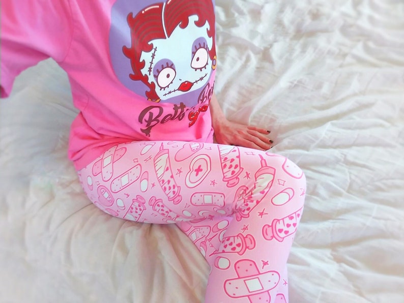 May include: Pink pajama set featuring a Betty Boop graphic on the shirt. The shirt is a solid pink color. The pajama pants have a light pink background with a pattern of medical-themed illustrations in a darker pink.