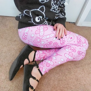 May include: Pink leggings with a repeating pattern of medical-themed illustrations, including bandages, syringes, and hearts. The leggings are paired with a black sweater featuring white graphic designs and black flats.