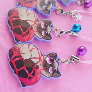May include: A purple charm with a cartoon cat tied up with pink rope. The charm has a gold ring and a silver chain attached to it.