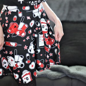 May include: A black skater skirt with a red and white print of cartoon teddy bears, bandages, pills, and hearts. The skirt has a pleated design and is made of a soft, stretchy fabric.