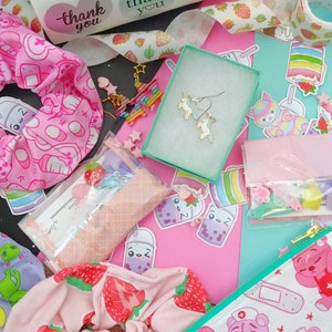 May include: A flat lay of a variety of items including a pink and white patterned scrunchie, a pair of unicorn earrings, a small pink pouch with a white bear design, and a variety of stickers with the text "thank you".