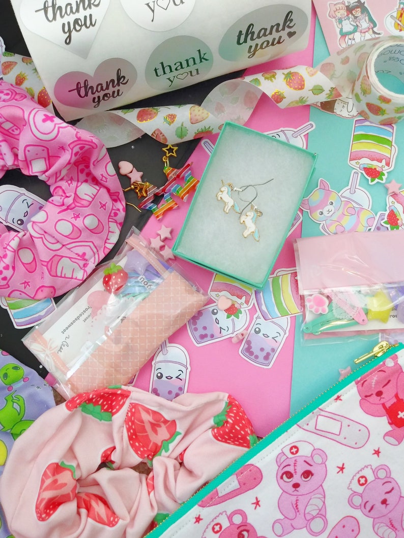 May include: A flat lay of a variety of items including a pink and white patterned scrunchie, a pair of unicorn earrings, a small pink pouch with a white bear design, and a variety of stickers with the text "thank you".
