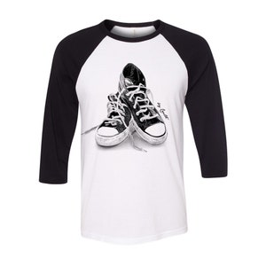May include: A white and black raglan baseball tee featuring a black and white illustration of a pair of sneakers. The sleeves are black, and the body is white. The graphic includes the text "by Corbet".