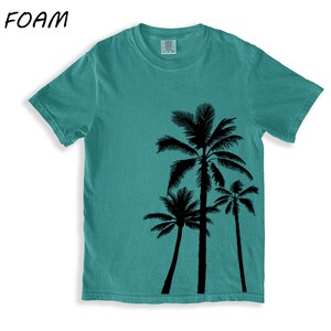 K&ouml;nnte beinhalten: Meergr&uuml;nes T-Shirt mit schwarzem Palmen-Motiv. Das T-Shirt hat einen Rundhalsausschnitt und kurze &Auml;rmel. Der Text "SEA FOAM" befindet sich in der oberen linken Ecke. Die Grafik zeigt drei unterschiedlich hohe Palmen.