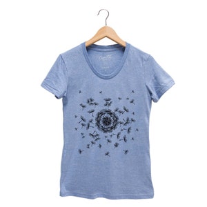 May include: A light blue t-shirt with a black dandelion design. The dandelion is in the center of the shirt and has seeds blowing in the wind. The shirt has a crew neck and short sleeves.