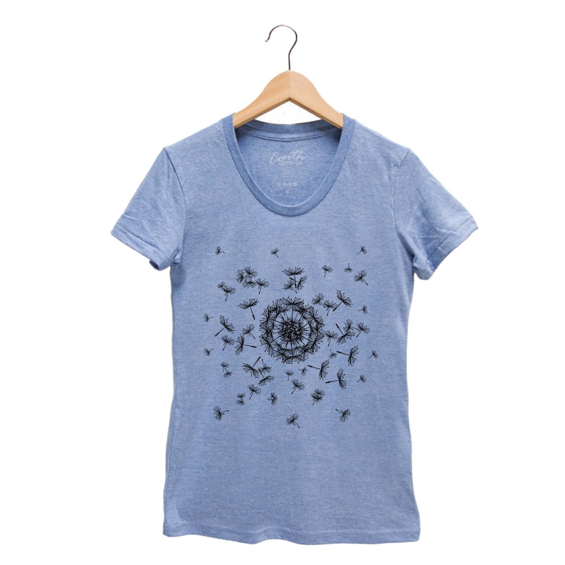 May include: A light blue t-shirt with a black dandelion design. The dandelion is in the center of the shirt and has seeds blowing in the wind. The shirt has a crew neck and short sleeves.