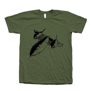 以下が含まれることがあります： 黒いSR-71ブラックバード機の白黒イラストが描かれた緑色のTシャツ。イラストには「by Scott」の署名があります。