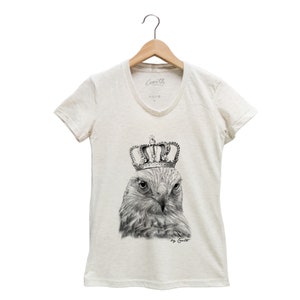 May include: A white t-shirt with a black and white illustration of a hawk wearing a gold crown. The text "by Coult" is printed below the illustration.