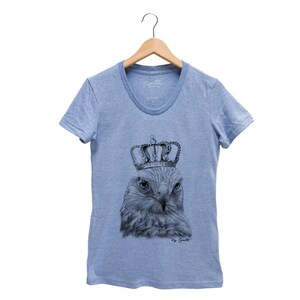 May include: A light blue t-shirt with a black and white illustration of a hawk wearing a crown. The text "by Caito" is printed below the illustration.