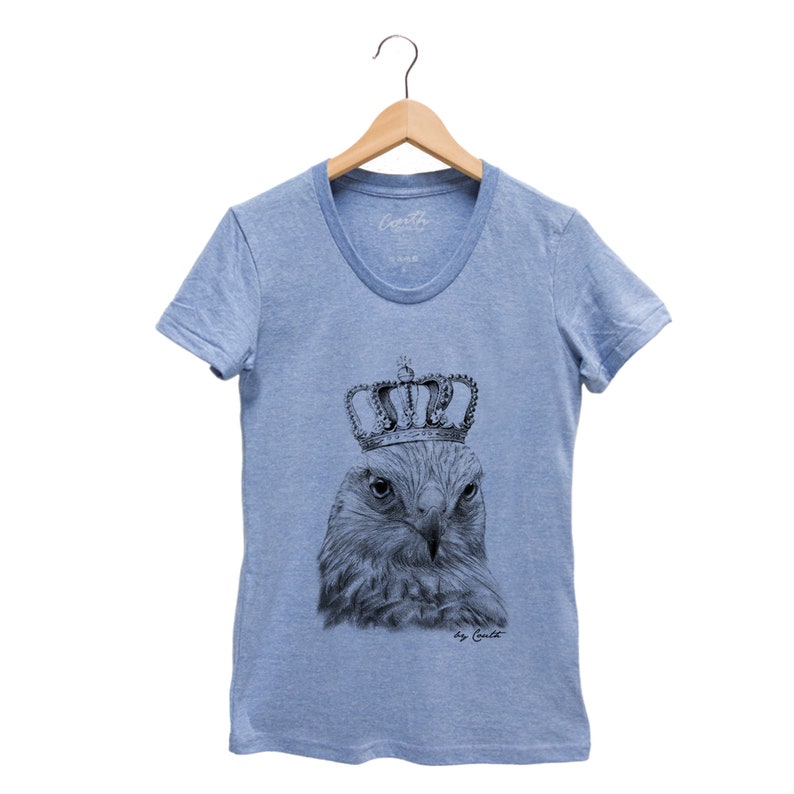 May include: A light blue t-shirt with a black and white illustration of a hawk wearing a crown. The text "by Caito" is printed below the illustration.