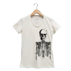 Puede incluir: Una camiseta blanca con un gr&aacute;fico de esqueleto en blanco y negro. El esqueleto est&aacute; de pie con los brazos a los lados. La camiseta tiene cuello en V y mangas cortas.