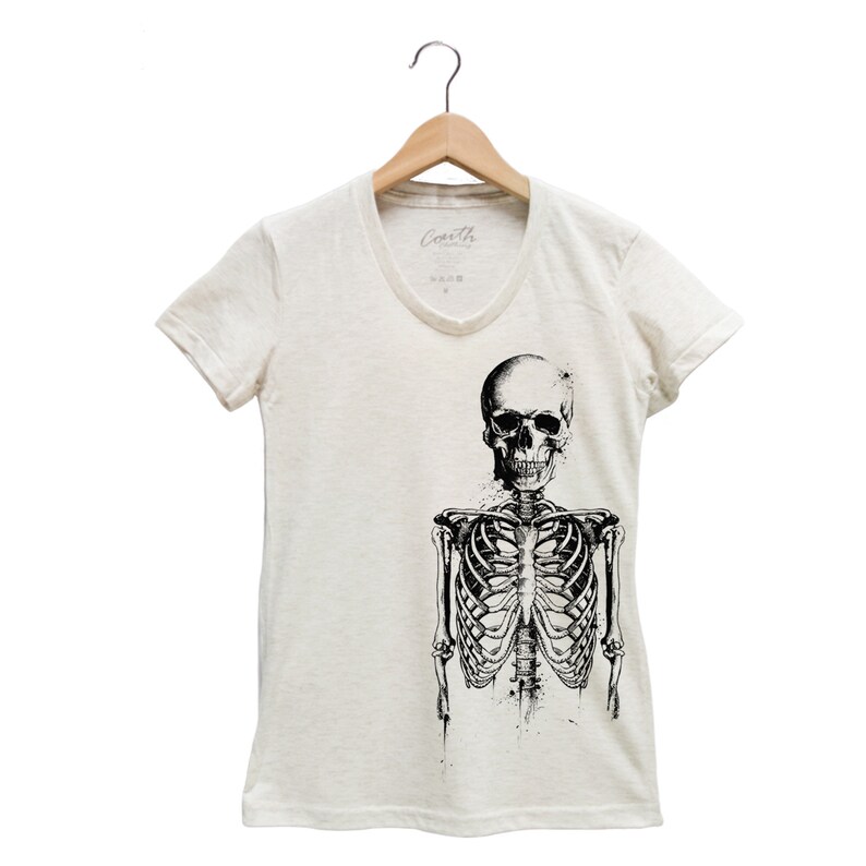 Puede incluir: Una camiseta blanca con un gr&aacute;fico de esqueleto en blanco y negro. El esqueleto est&aacute; de pie con los brazos a los lados. La camiseta tiene cuello en V y mangas cortas.