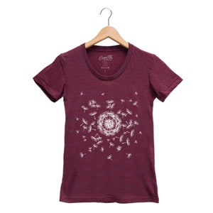 May include: A maroon t-shirt with a white dandelion design. The dandelion is in the center of the shirt and has seeds blowing in the wind. The shirt has a crew neck and short sleeves.