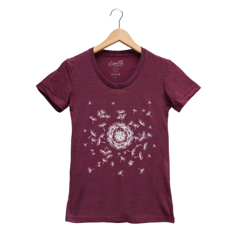 May include: A maroon t-shirt with a white dandelion design. The dandelion is in the center of the shirt and has seeds blowing in the wind. The shirt has a crew neck and short sleeves.