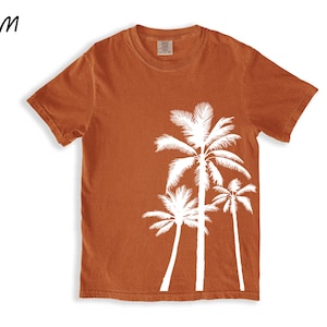 K&ouml;nnte beinhalten: Ein rostfarbenes T-Shirt mit einem wei&szlig;en Palmen-Grafikdesign. Das Design zeigt drei Palmen. Das Shirt hat einen Rundhalsausschnitt und kurze &Auml;rmel. Das Wort "YAM" befindet sich in der oberen linken Ecke.