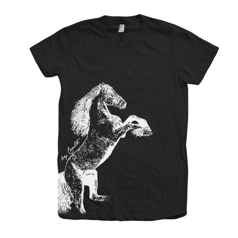 Op de afbeelding: Zwart T-shirt met een wit silhouet van een paard dat op zijn achterpoten staat. Het paard is gedetailleerd met een manen en staart. De tekst "by Cynthia" is zichtbaar aan de linkerkant van de afbeelding.