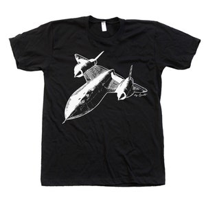 May include: Black t-shirt featuring a white illustration of a jet plane. The graphic depicts the aircraft in flight, with detailed wings and body. The text "by Conalt" is printed in the lower right corner of the graphic.