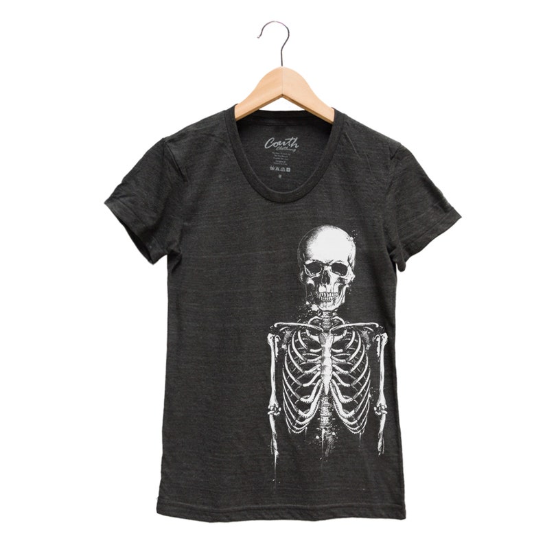 Puede incluir: Una camiseta gris oscuro con un gr&aacute;fico de esqueleto blanco. El esqueleto est&aacute; de pie con los brazos a los lados. El cr&aacute;neo est&aacute; mirando hacia adelante. La camiseta tiene un cuello redondo y mangas cortas.