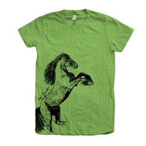 Op de afbeelding: Een groen T-shirt met een zwart-witte illustratie van een paard dat op zijn achterpoten staat. De manen van het paard stromen en de illustratie is gedetailleerd. De tekst "Roy Cavett" is in cursief geschreven op de zijkant van het T-shirt.