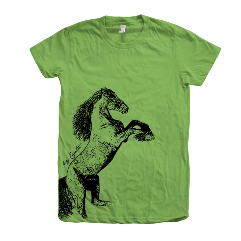 Op de afbeelding: Een groen T-shirt met een zwart-witte illustratie van een paard dat op zijn achterpoten staat. De manen van het paard stromen en de illustratie is gedetailleerd. De tekst "Roy Cavett" is in cursief geschreven op de zijkant van het T-shirt.